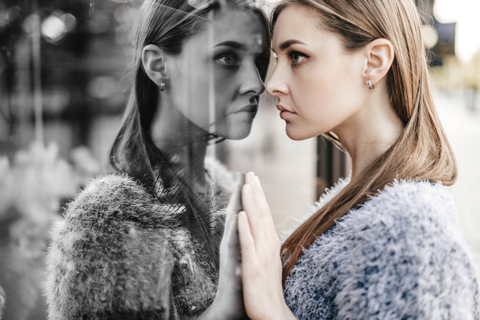 Eine Junge Frau lehnt frontal an einer Scheibe und betrachtet ihr schwarz-weißes Spiegelbild.