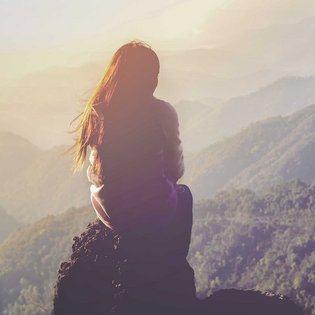 Eine Frau sitzt auf einem Berg und schaut in das bewaldete Tal hinab.
